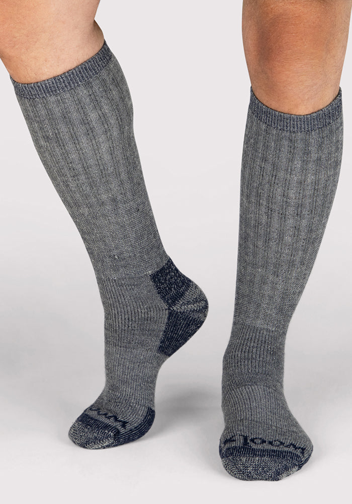 A person wearing Woolx Mens Extreme Over The Calf Socks in Navy Carbon, featuring heavyweight Merino wool with ribbed fabric and dark gray accents on the toes, heels, and soles, stands on a light background.