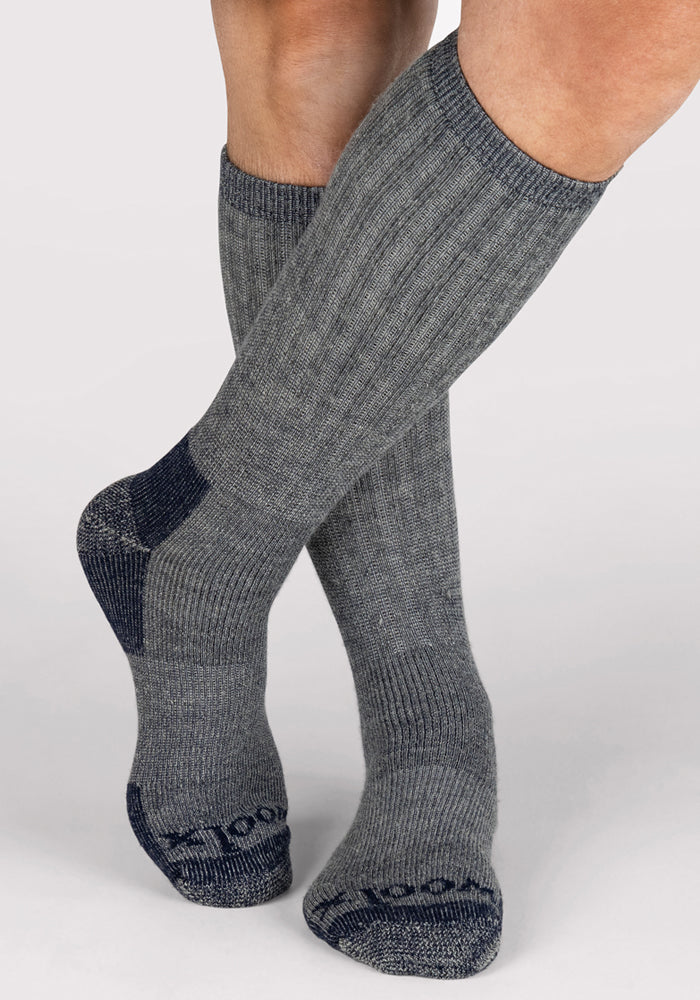 A pair of feet wearing Woolx Mens Extreme Over The Calf Socks in Navy Carbon, featuring tall, ribbed design with darker reinforced heels, toes, and soles, stands against a plain light background.