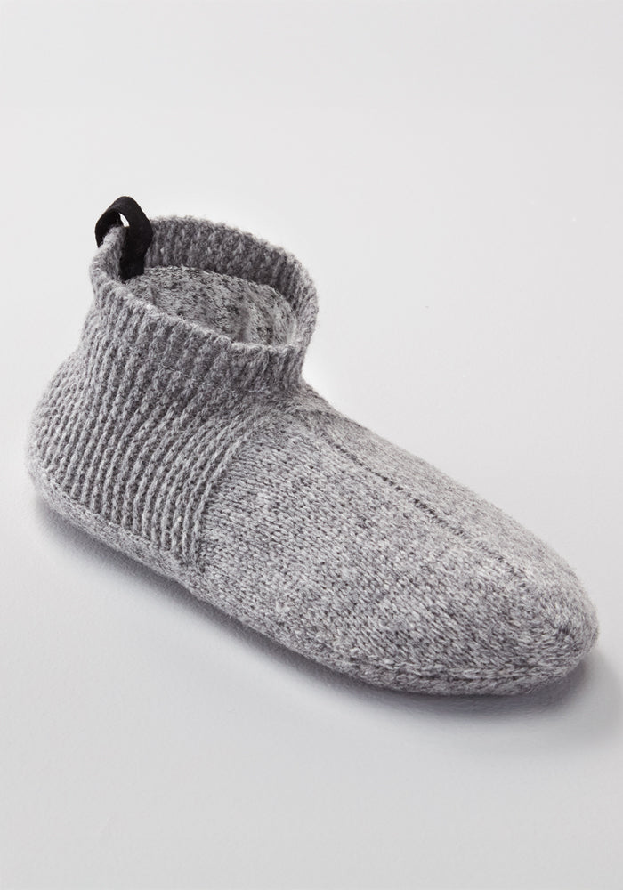 The Woolx Mens Gripper Slippers in Gentle Grey feature a single gray knitted merino wool design with a ribbed ankle cuff and a black pull tab at the back, shown on a light surface.