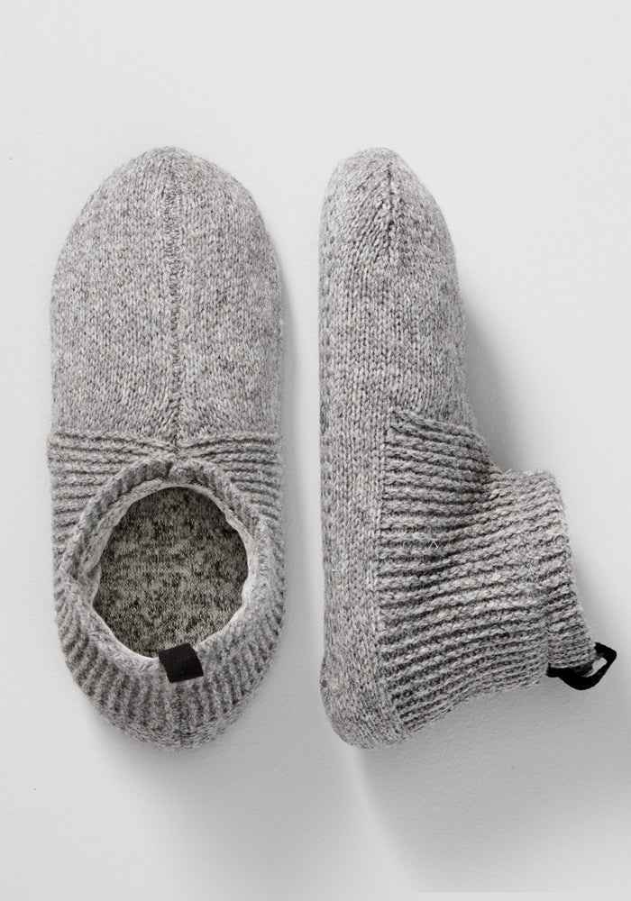 A pair of Woolx Mens Gripper Slippers in Gentle Grey; one is shown top-down to display the opening, while the other is on its side highlighting the ribbed ankle cuff and textured knit fabric.