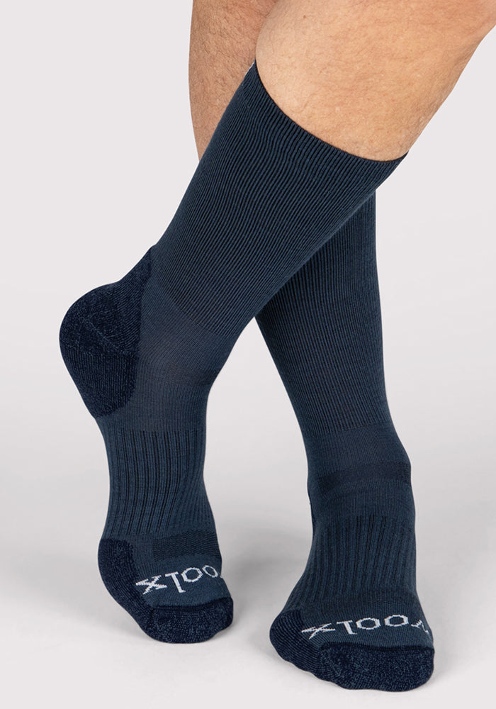 A person wearing Woolx Mens Harbor Crew Sock - Evening Tide, dark blue Merino wool with ribbing and reinforced heels/toes, stands with one foot crossed in front; "1001" appears on the toes.