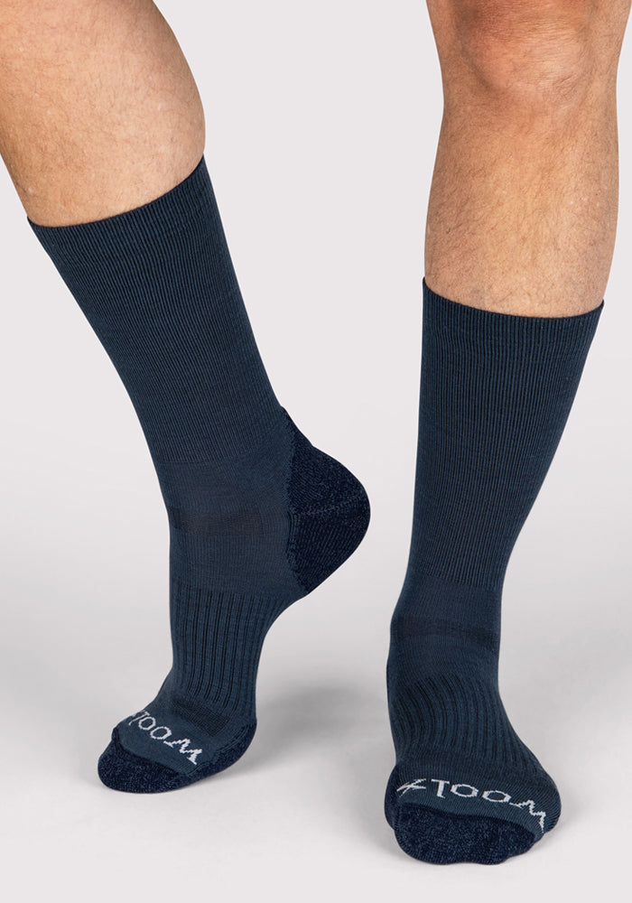 A person wears Woolx Mens Harbor Crew Sock - Evening Tide, featuring navy blue, odor-resistant fabric with ribbed texture and white text on the soles, while standing on a light gray surface.