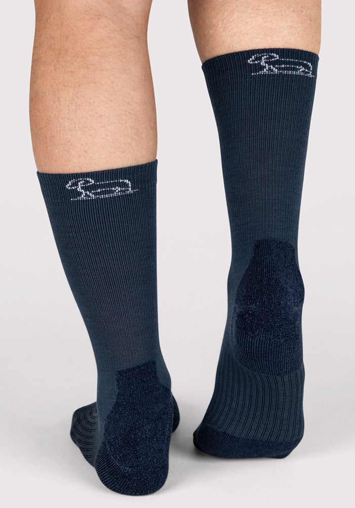 A person seen from behind, wearing Woolx Mens Harbor Crew Sock in Evening Tide—navy blue, ribbed, moisture-wicking socks with reinforced heels and a white embroidered logo near the top—against a plain light background.