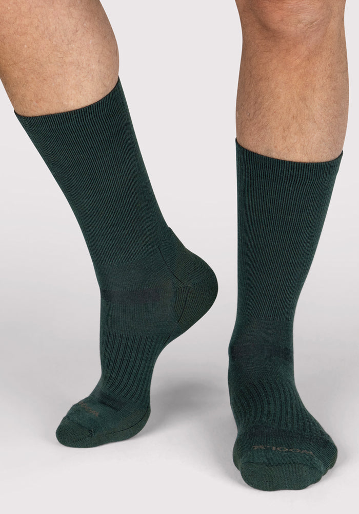A person wears Woolx Mens Harbor Crew Sock in Forest, standing against a light gray background with one foot flat and the other raised onto its toes.