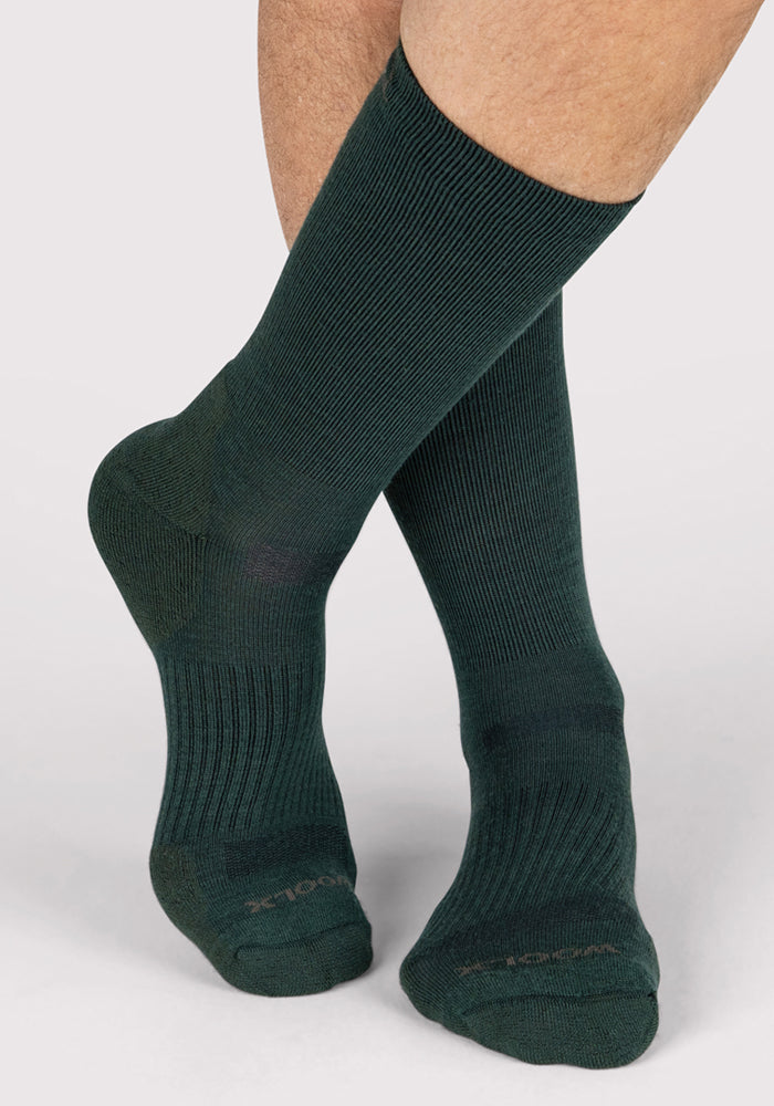 A close-up of a person's lower legs and feet wearing Woolx Mens Harbor Crew Sock in Forest, made from moisture-wicking Merino wool. The person stands with one foot crossed in front against a plain light background.