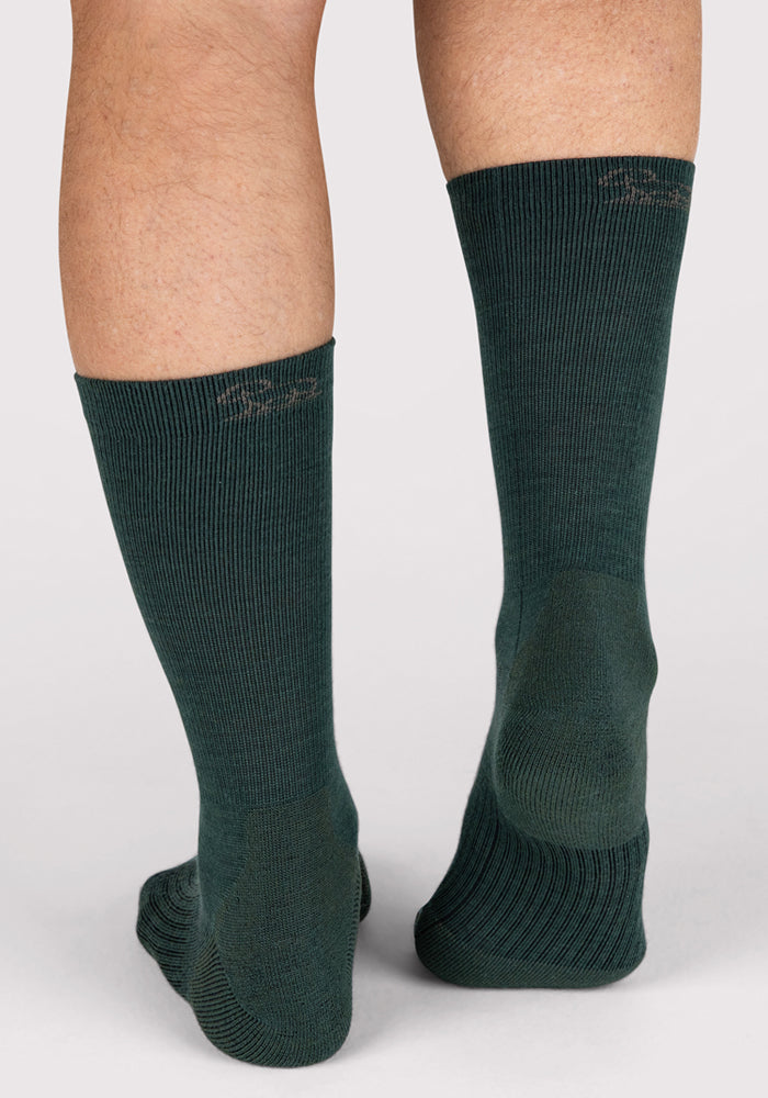 A person wearing Woolx Mens Harbor Crew Sock in Forest, made of moisture-wicking Merino wool, stands on a light, neutral background, displaying the back and sides of their lower legs and feet.