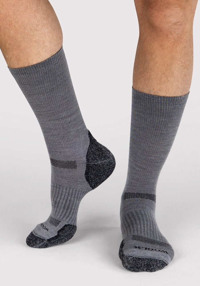 A person wears Mens Harbor Crew Socks - Grey by Woolx, featuring moisture-wicking fabric, ribbed cuffs, and dark gray heel, toe, and arch accents. Subtle horizontal lines enhance comfort. They stand on a light background.