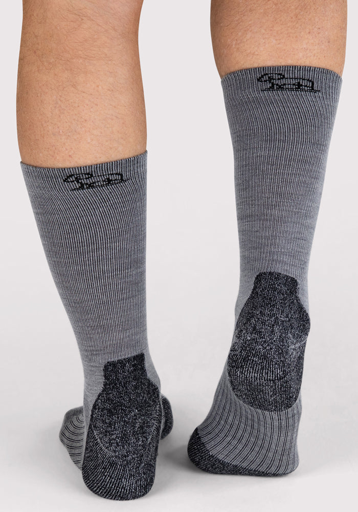 A close-up of a person wearing Woolx Mens Harbor Crew Socks in grey, featuring ribbed moisture-wicking fabric, black reinforced heels, and a small black logo near the top, standing on a light background.