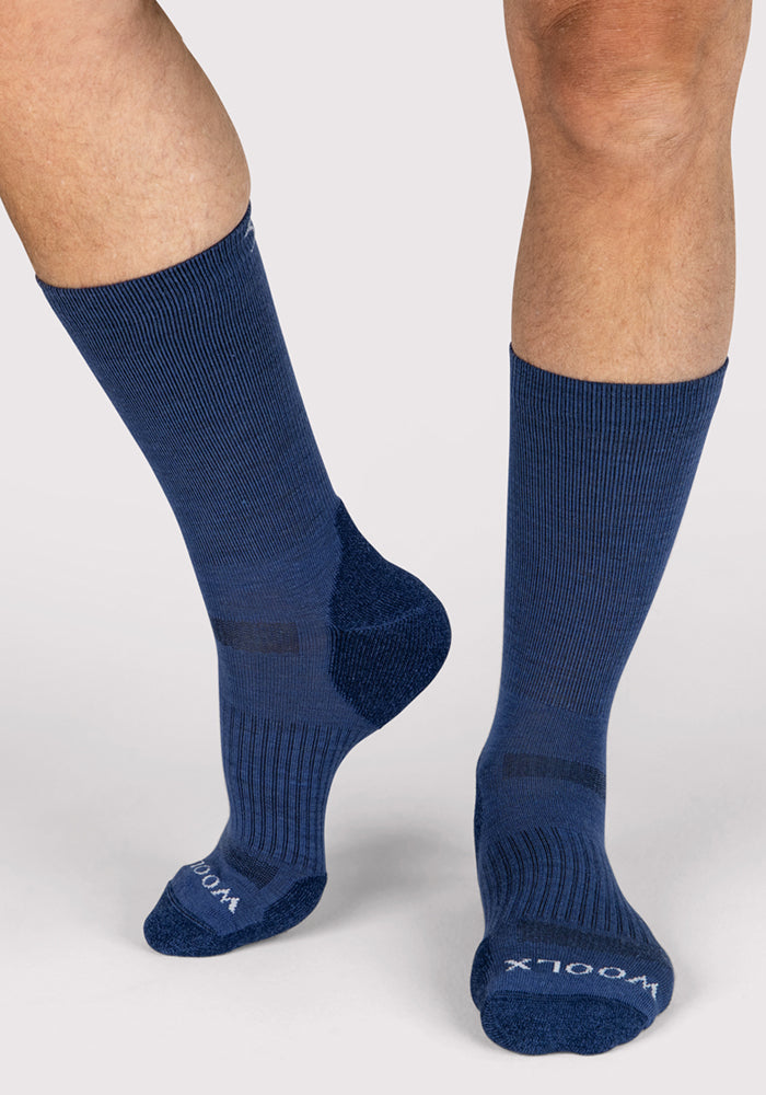 A person wears Woolx Mens Harbor Crew Socks in Sailor Blue, featuring ribbed Merino wool and "WOOLX" on the toes, standing on a light gray background.