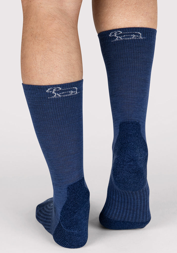 A close-up of a person wearing Woolx Mens Harbor Crew Socks in Sailor Blue, featuring moisture-wicking fabric and a white logo near the top back, standing on a neutral surface.