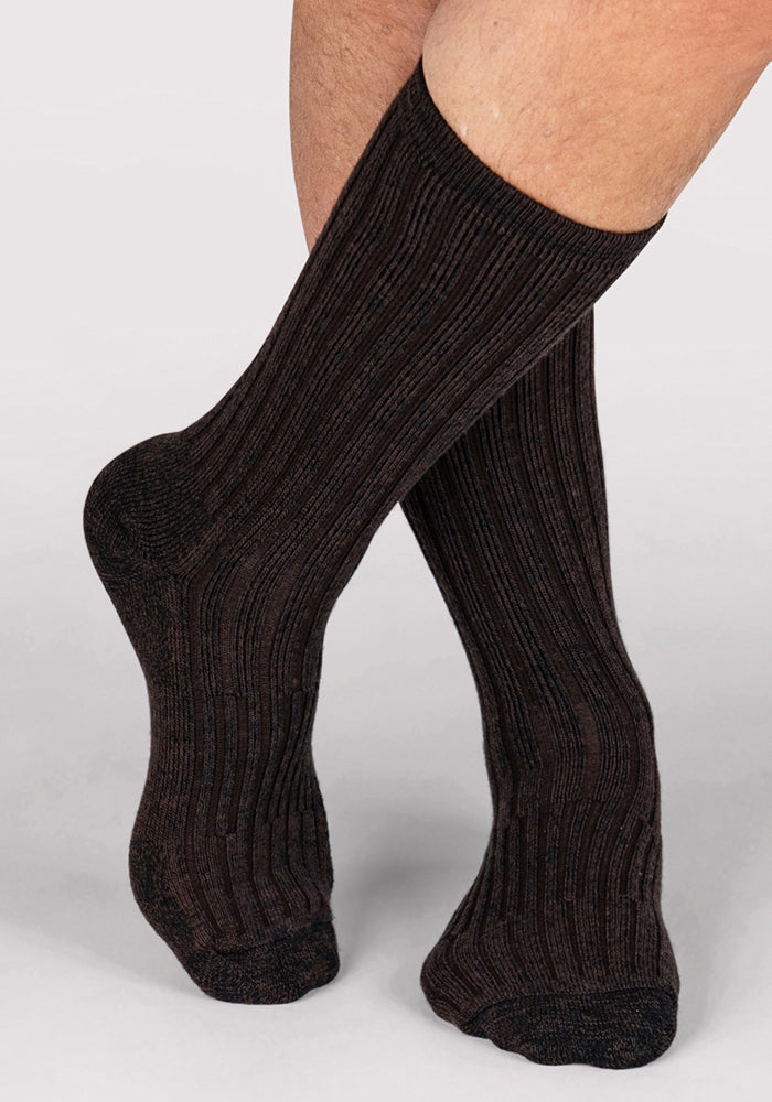 A person wearing Woolx Mens Joey Crew Socks in Dark Espresso stands with one foot crossed over the other against a light gray background.