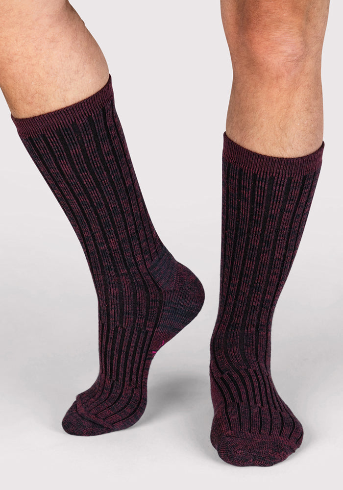 A person wears Woolx Mens Joey Crew Socks in Warm Violet, featuring a dark purple and black ribbed pattern, while standing on a light background.