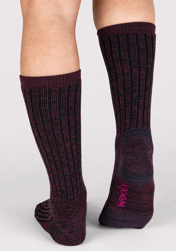 A person wearing Woolx Mens Joey Crew Socks in Warm Violet—dark, ribbed with pink sole lettering—stands on a light gray background. Only their lower legs and feet are visible.