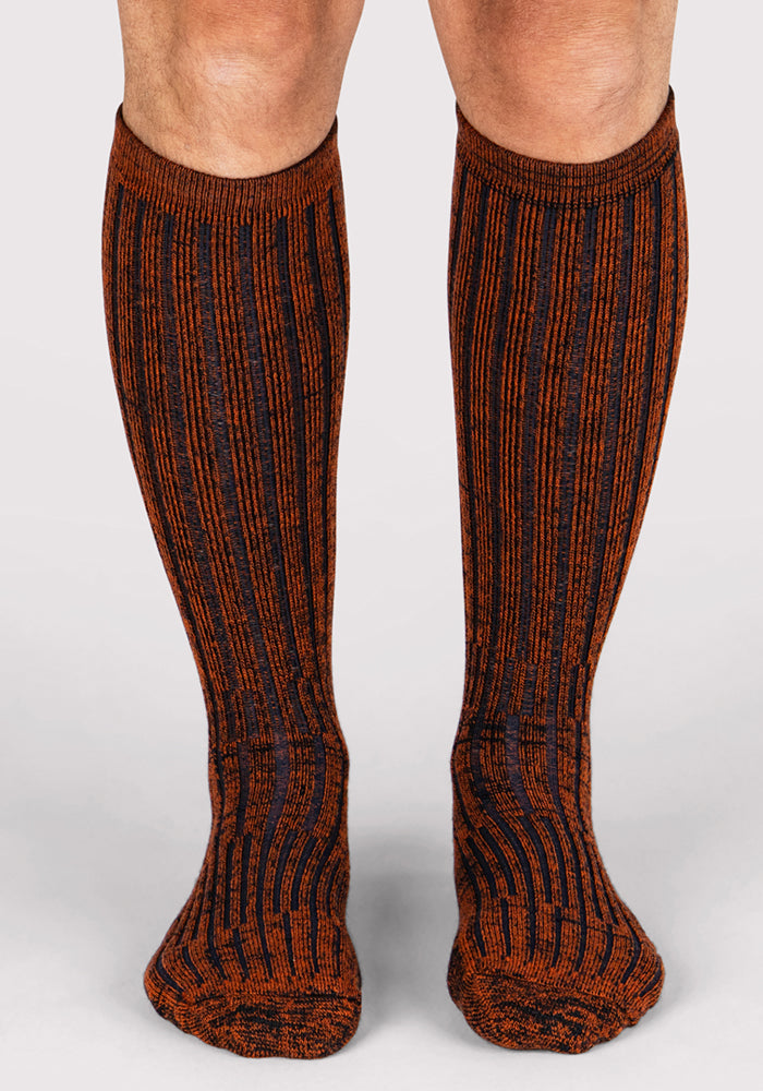A front view shows legs wearing Woolx Mens Joey Mid Calf Socks in Cinnamon Spice, featuring moisture-wicking fabric and vertical orange and black stripes, set against a plain light background.