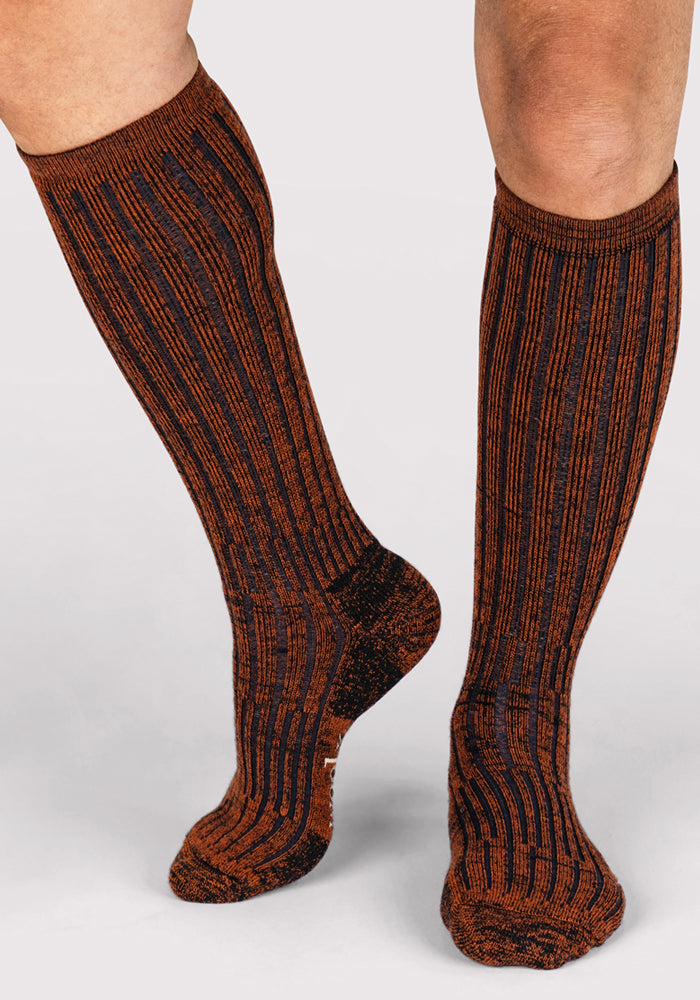 A person wearing Woolx Mens Joey Mid Calf Socks in Cinnamon Spice, featuring a vertical ribbed pattern in brown and black, stands on a light gray background.
