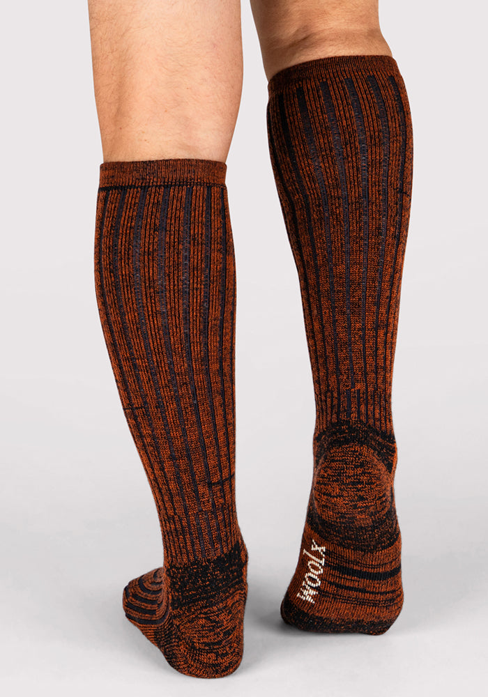 A person wears Woolx Mens Joey Mid Calf Socks in Cinnamon Spice—brown and black striped Merino wool socks with "WOOL" in white on the sole, standing on a light gray background. These moisture-wicking socks ensure all-day comfort.