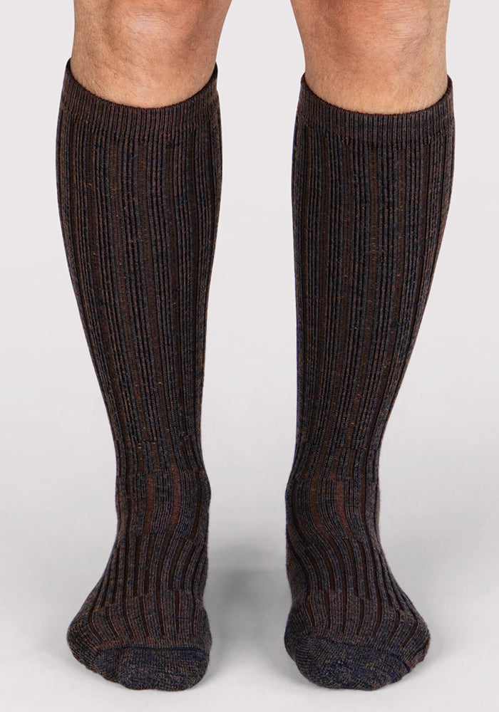 A person wears Woolx Mens Joey Mid Calf Socks in Dark Espresso, standing on a light background with only their lower legs and feet visible.