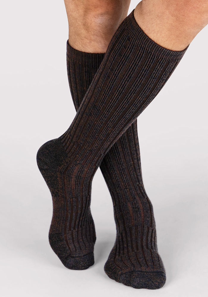 A person stands with one foot crossed in front of the other against a plain background, wearing Woolx Mens Joey Mid Calf Socks in Dark Espresso.
