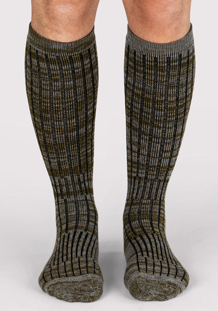 A person wearing Woolx Mens Joey Mid Calf Socks - Dark Forest Marled, featuring a ribbed texture in gray and olive green, stands on a plain light background.