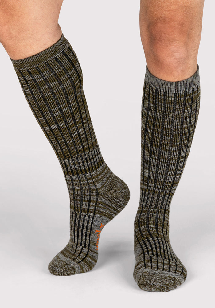 A person stands on a light gray background wearing Woolx Mens Joey Mid Calf Socks in Dark Forest Marled, featuring a ribbed merino wool knit with green and gray plaid, black vertical stripes, and orange text on the sole.