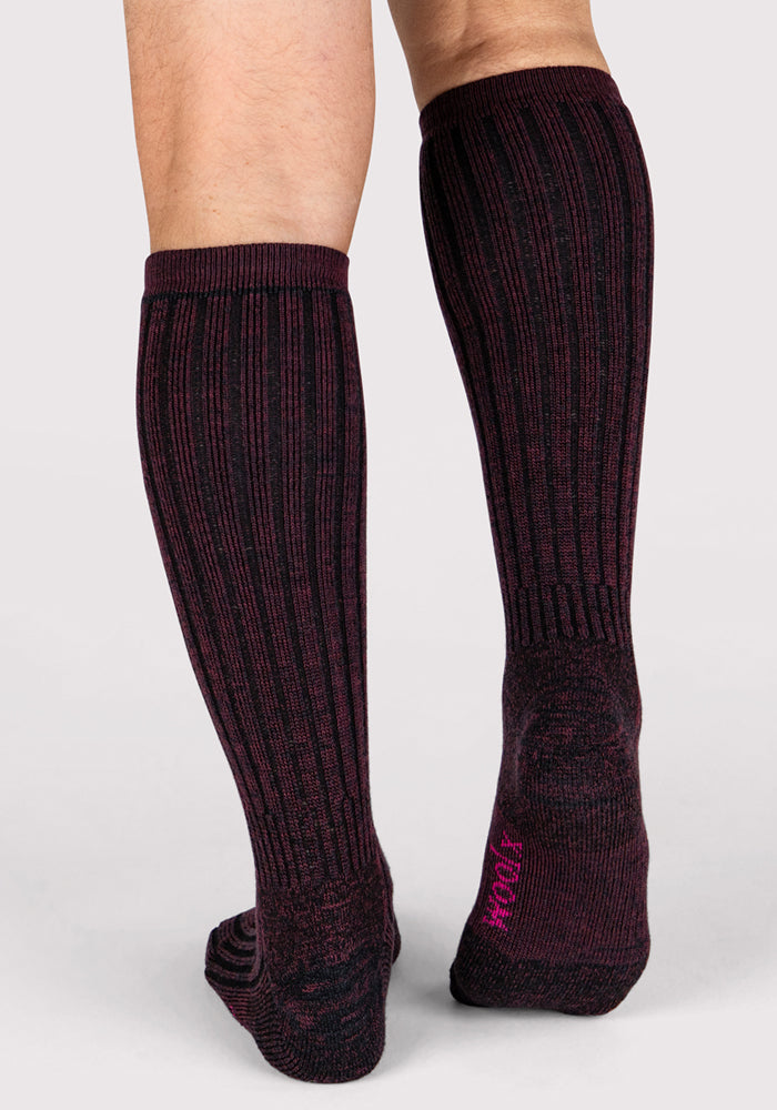 A person stands on a light background wearing Woolx Mens Joey Mid Calf Socks in Warm Violet, featuring a subtle maroon and black pattern with pink text on the moisture-wicking sole of the right foot.