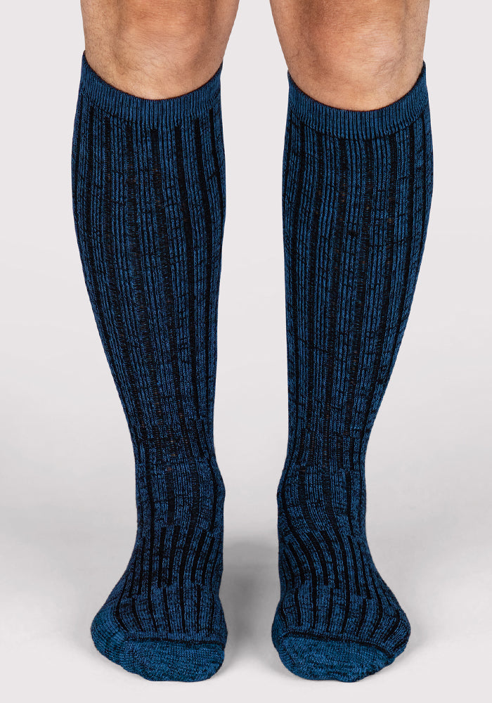 A person stands on a light gray surface wearing Woolx Mens Joey Mid Calf Socks in Steel Blue, featuring a ribbed design with blue and black vertical stripes.