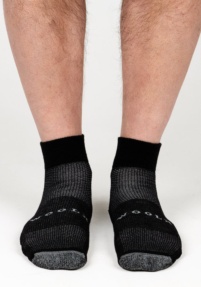 A close-up of a person wearing Woolx Mens Rambler Ankle Sock in Black Pepper, showcasing the black and gray patterned Merino wool for excellent moisture management, while standing on a white background.