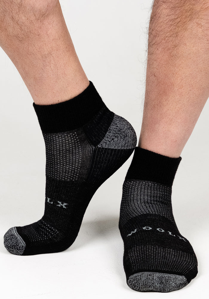 Close-up of a person’s lower legs wearing Woolx Mens Rambler Ankle Sock - Black Pepper, featuring textured patterns and moisture-managing Merino wool, with text on the top, standing on a white background.