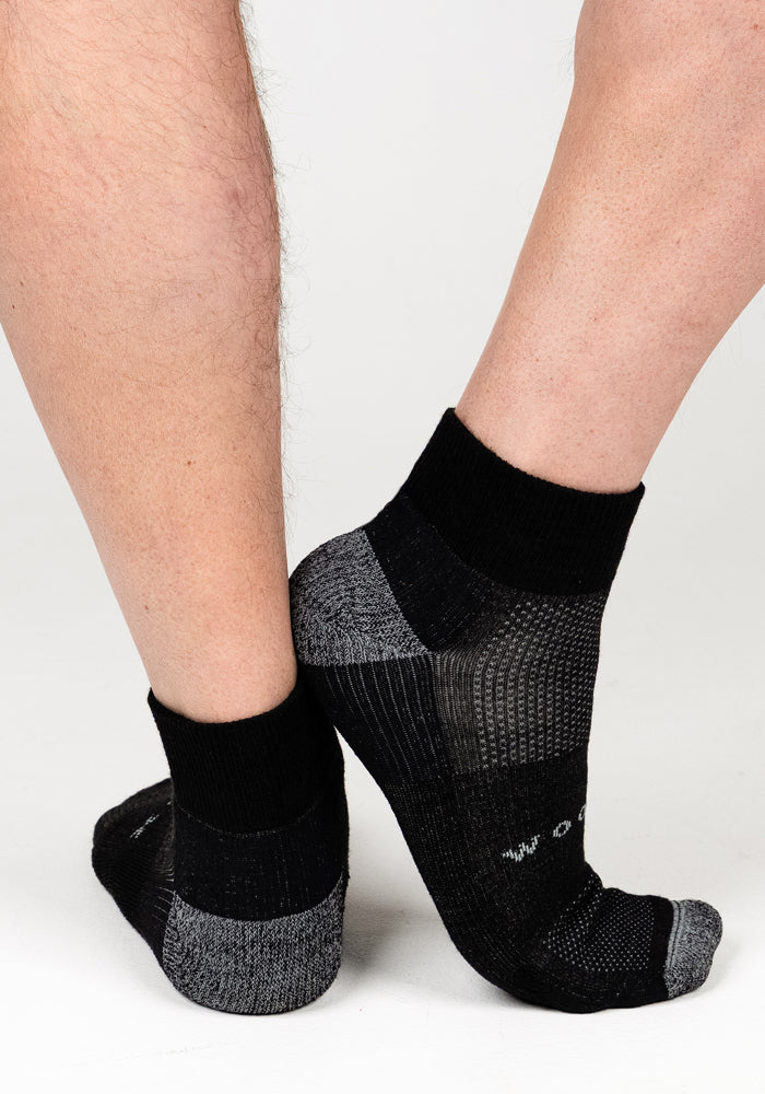 Two legs wearing Woolx Mens Rambler Ankle Sock - Black Pepper are crossed at the ankles against a white background. The socks have a textured pattern on the sides and toes for all-day comfort.