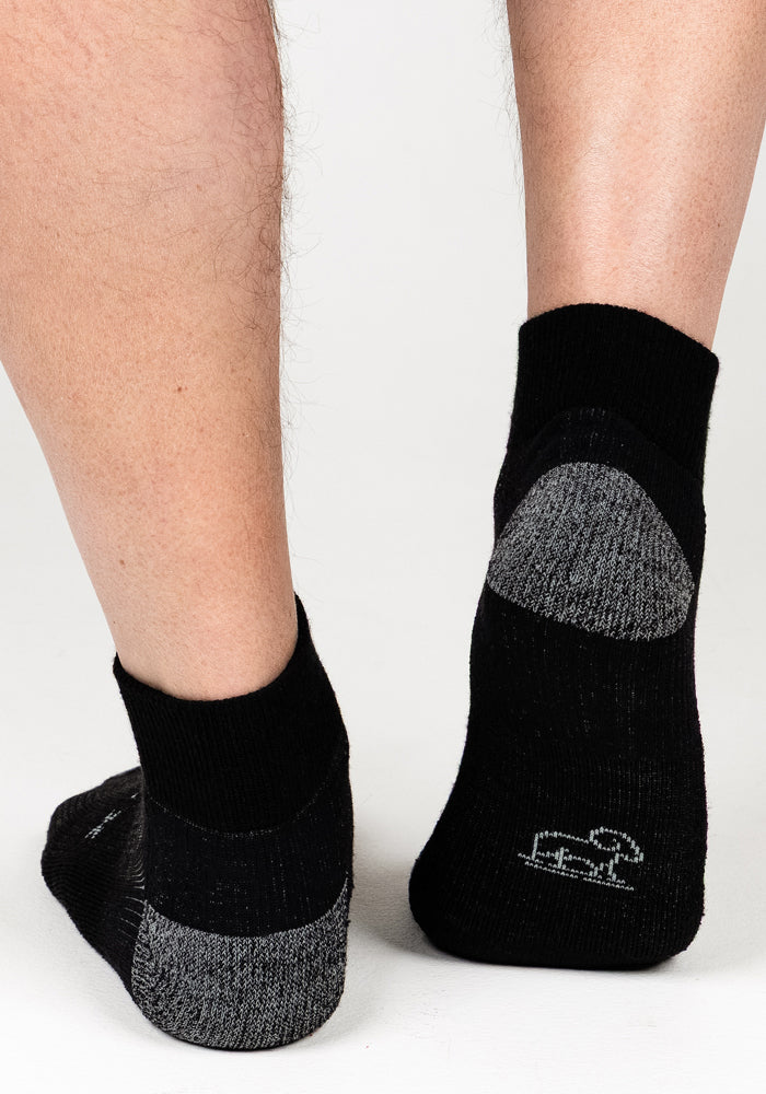 A close-up of two feet standing on a white surface showcases the Woolx Mens Rambler Ankle Sock in Black Pepper, featuring gray accents on the heels and toes and offering superior moisture management.