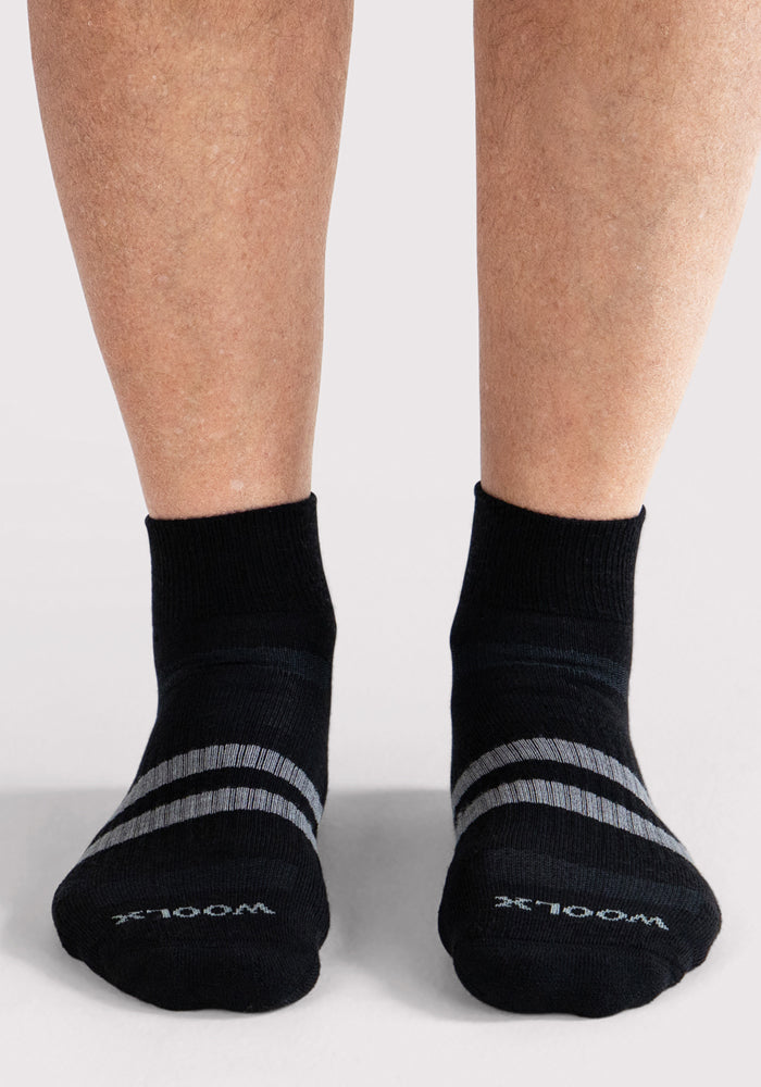 A person stands on a light background wearing Mens Rambler Ankle Sock • Final Sale - Black by Woolx. These black Merino wool ankle socks feature gray stripes and the "WOOLX" logo near the toes, highlighting their moisture-wicking design.