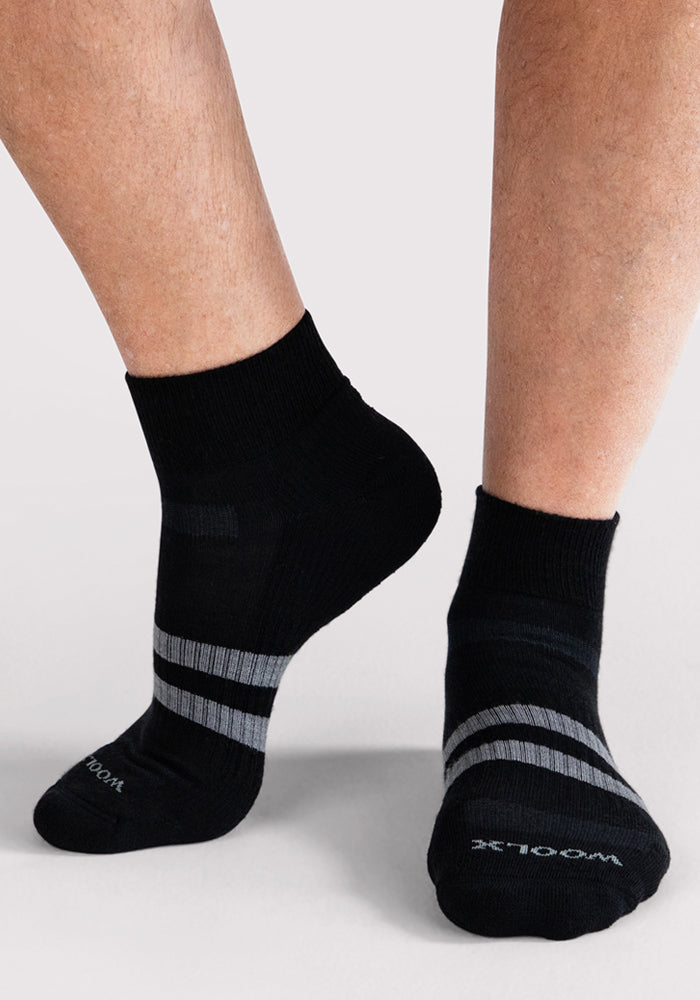A person wears Woolx Mens Rambler Ankle Sock • Final Sale - Black, featuring two gray stripes near the toes on a light gray surface—ideal for all-day moisture-wicking comfort.