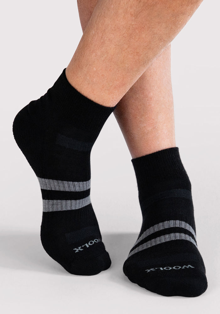 A person wearing Woolx Mens Rambler Ankle Sock • Final Sale - Black, featuring gray stripes and moisture-wicking fabric, stands with one foot crossed over the other against a light gray background.