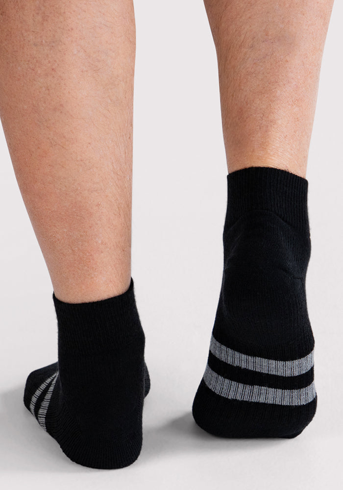 A close-up of a person's lower legs and feet wearing Woolx Mens Rambler Ankle Sock • Final Sale - Black, featuring two gray stripes near the cuffs, standing on a light-colored surface.