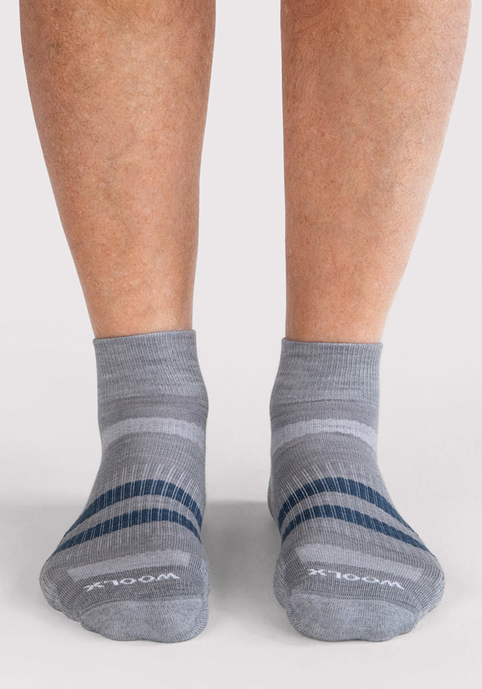 A pair of feet wearing Woolx Mens Rambler Ankle Sock - Grey, featuring dark gray and blue stripes and moisture-wicking fabric, stands on a light-colored surface.