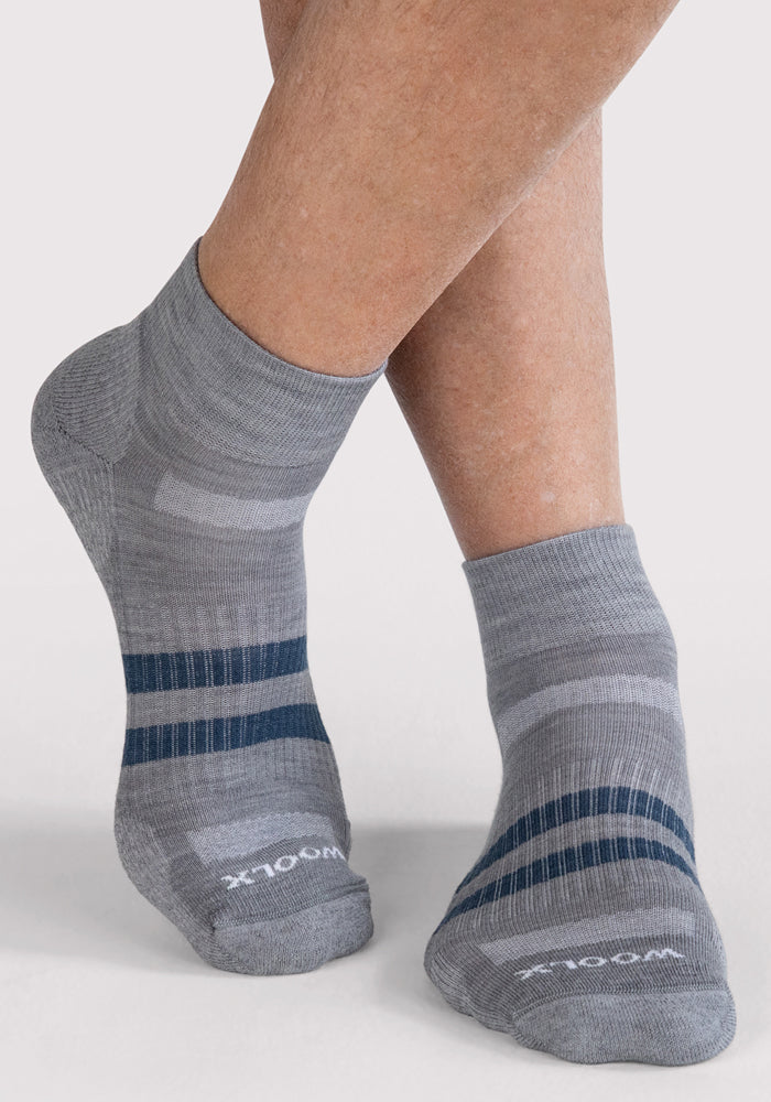 Close-up of a person wearing Woolx Mens Rambler Ankle Sock in grey with blue and light gray stripes. Moisture-wicking socks, "WOOL" on the toe, feet crossed at the ankles on a plain white background.