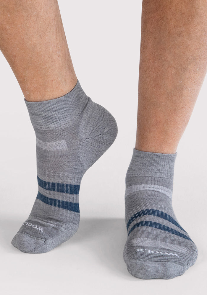 A person wearing Woolx Mens Rambler Ankle Sock - Grey, featuring dark blue and light gray stripes, ribbed cuffs, and moisture-wicking snug fit, stands on a light surface.