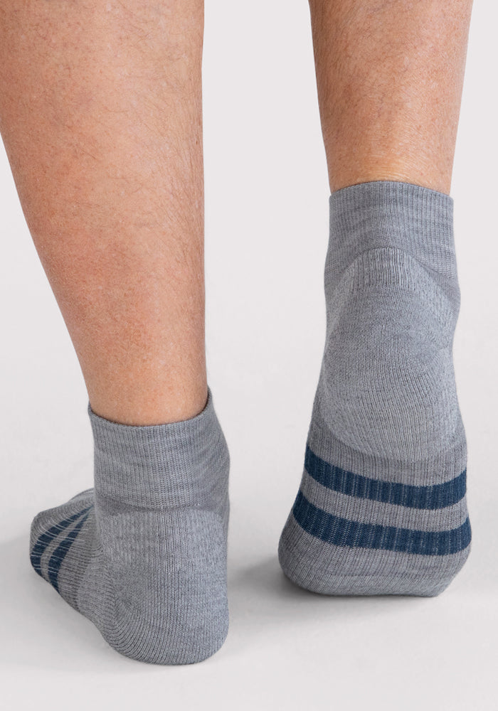 A close-up of two feet wearing Woolx Mens Rambler Ankle Sock - Grey, featuring moisture-wicking fabric and two dark blue stripes near the cuffs, shown from the back on a white background.