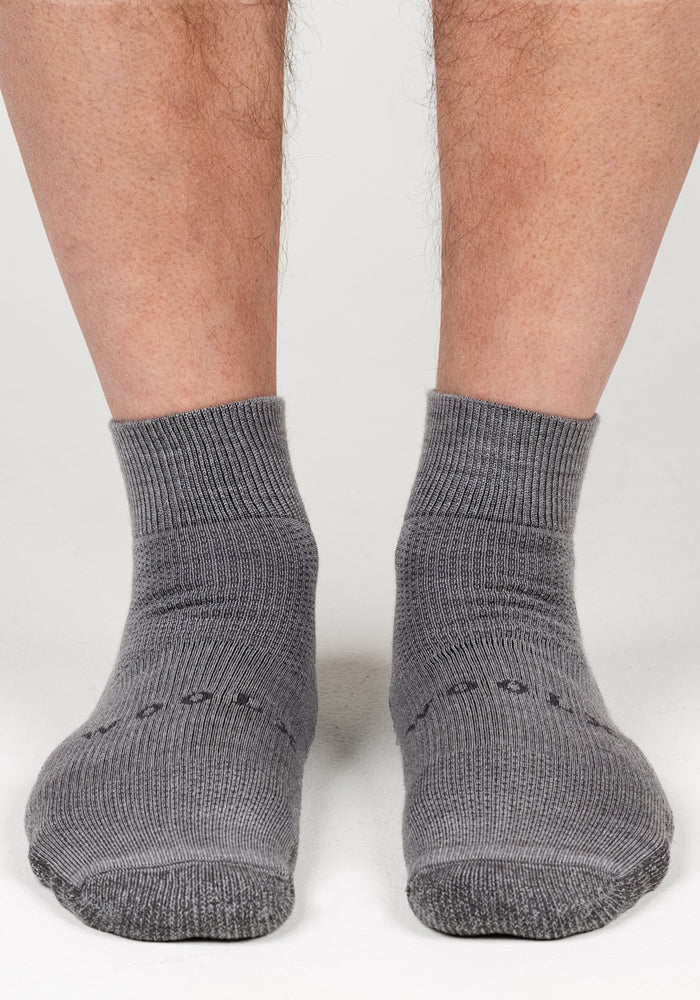 A pair of feet in Woolx Mens Rambler Ankle Sock - Pebble stands on a light surface, showcasing the socks’ moisture management. Only the lower legs and feet are visible.
