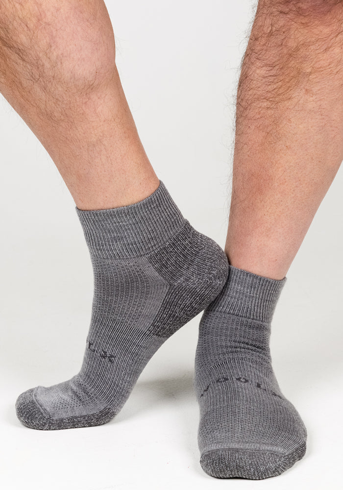 A close-up of a person's lower legs and feet wearing Woolx Mens Rambler Ankle Sock - Pebble shows the ribbed gray Merino wool with darker heel and toe accents, highlighted against a white background for a premium, moisture-managing look.