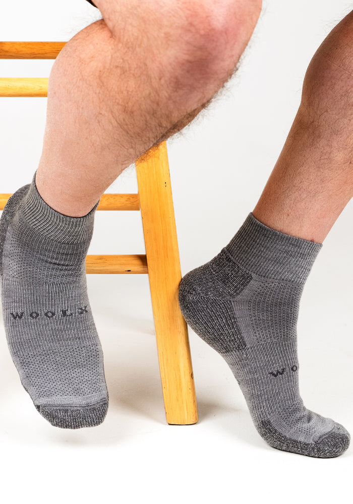 A person wearing Woolx Mens Rambler Ankle Sock in Pebble, featuring a textured pattern and moisture management, sits with one foot on the floor and the other on a wooden stool. "Woolx" is visible on the side of the socks.