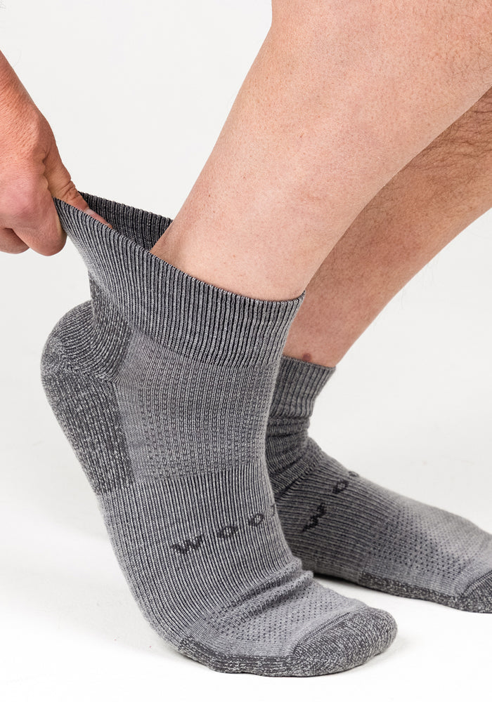 A person wearing Woolx Mens Rambler Ankle Sock in Pebble adjusts one sock upwards for a comfortable fit against a plain white background.