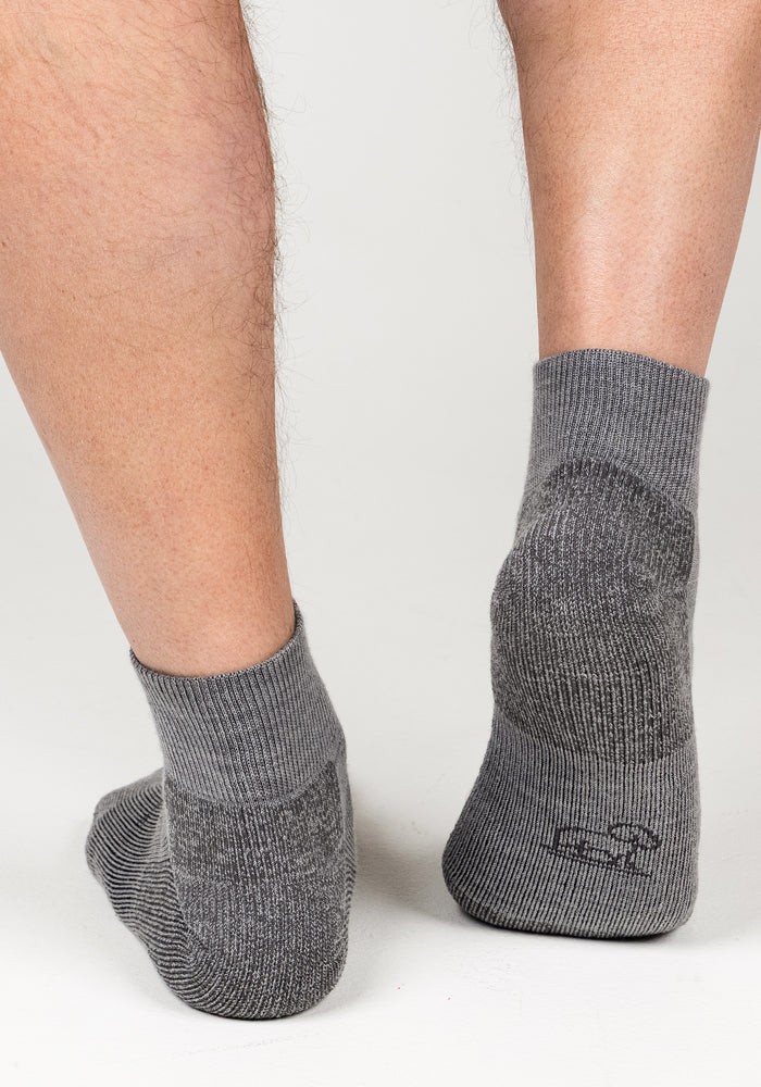 Close-up of a person's lower legs and feet wearing Woolx Mens Rambler Ankle Sock - Pebble, featuring a textured heel, moisture management, and a small logo, standing on a white surface.