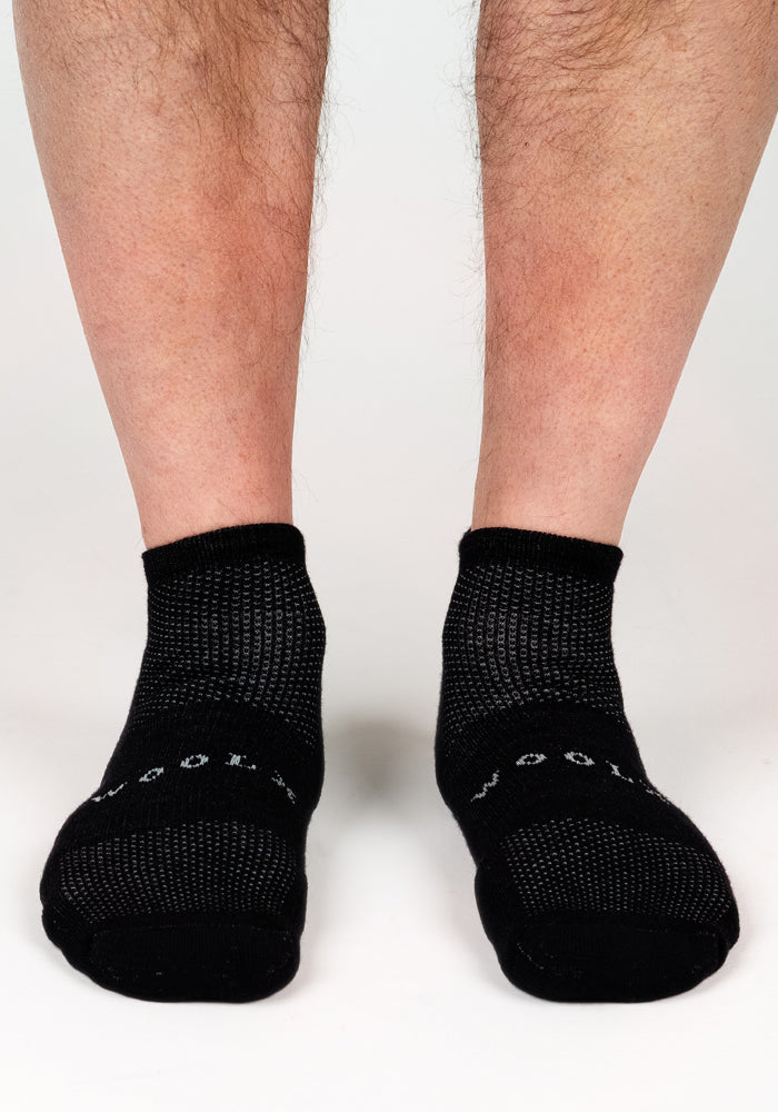 A close-up of lower legs and feet wearing Woolx Mens Rowan No Show Socks in Black Pepper. The textured Merino wool socks feature "WOOL" on the toes and offer odor-resistant comfort, displayed against a white background.