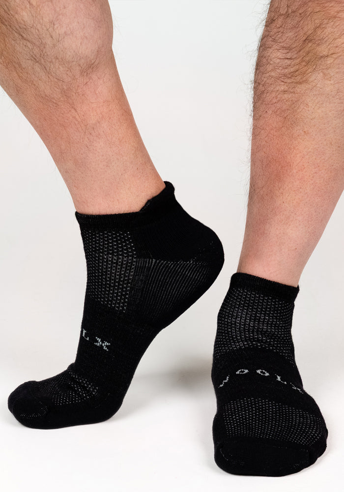A close-up of two feet wearing Woolx Mens Rowan No Show Socks in Black Pepper with a textured pattern and white lettering, set against a plain white background.