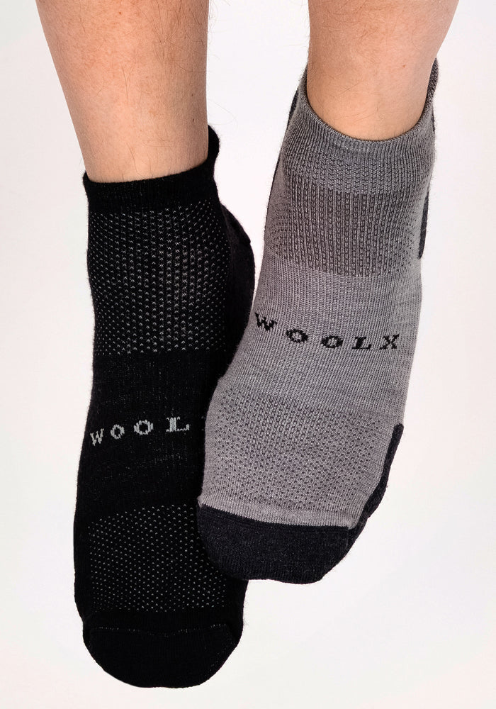 Two feet wear Woolx Mens Rowan No Show Socks—one black with white "WOOLX," one gray with black "WOOLX." These odor-resistant, cushioned Merino wool socks have textured patterns and an ankle height for comfort.