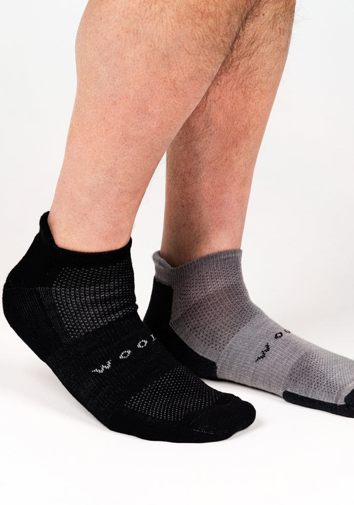 A person wears mismatched Mens Rowan No Show Socks by Woolx—one Black Pepper sock on the left foot and one gray patterned sock on the right—shown against a plain white background.
