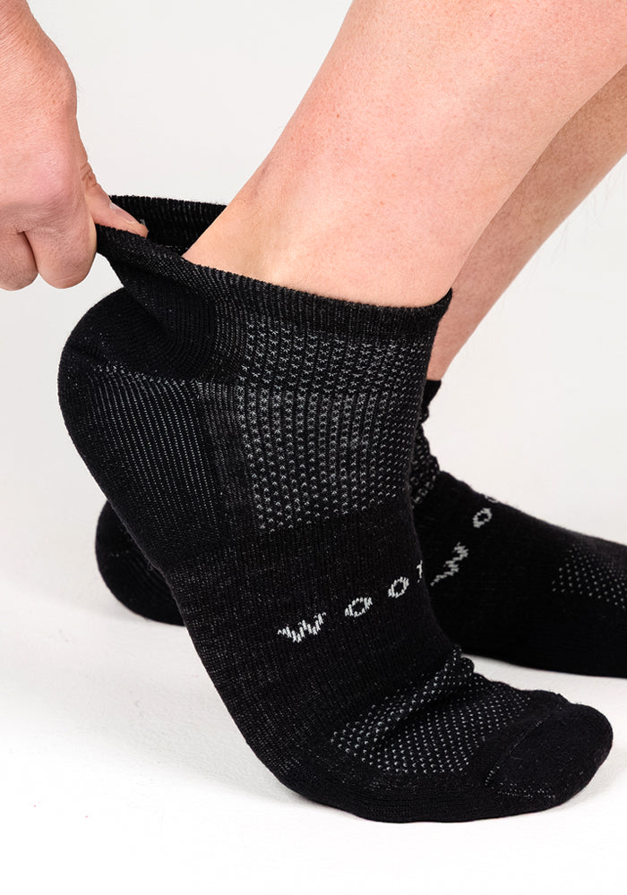 A person is pulling on a pair of Woolx Mens Rowan No Show Socks in Black Pepper, featuring a textured pattern and white lettering near the toes, against a plain white background.