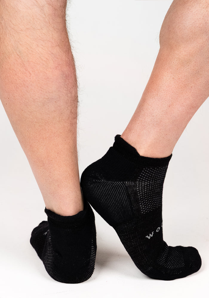 Close-up of two legs—one hairy, one smooth—both wearing Woolx Mens Rowan No Show Socks in Black Pepper, featuring moisture-wicking fabric and textured patterns, set against a plain white background.
