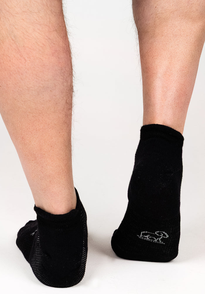 A person’s lower legs and feet wearing Woolx Mens Rowan No Show Socks in Black Pepper stand on a white background, heels and back of the legs visible. The right sock features a small white logo near the heel.